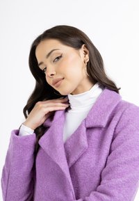 Lilla uldfrakke med nøglelapel, struktureret overflade og lange ærmer. Styled over en hvid turtleneck skjorte, med sølvfargede hoop øreringe.