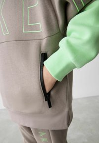 Hellbraunes Sweatshirt mit grünen Akzenten und Text, ausgestattet mit einer seitlichen Reißverschlusstasche und gerippten Bündchen. Weicher Stoff.
