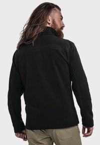 Zwarte fleec-jack met een hoge kraag en lange mouwen, met een gladde textuur en modern ontwerp, gedragen over lichtgekleurde broeken.
