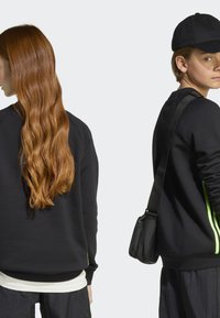 Sweatshirt preto com mangas longas, apresentando riscas verdes néon. Acompanhado de um boné preto e uma bolsa de ombro. Textura suave, ajuste descontraído.