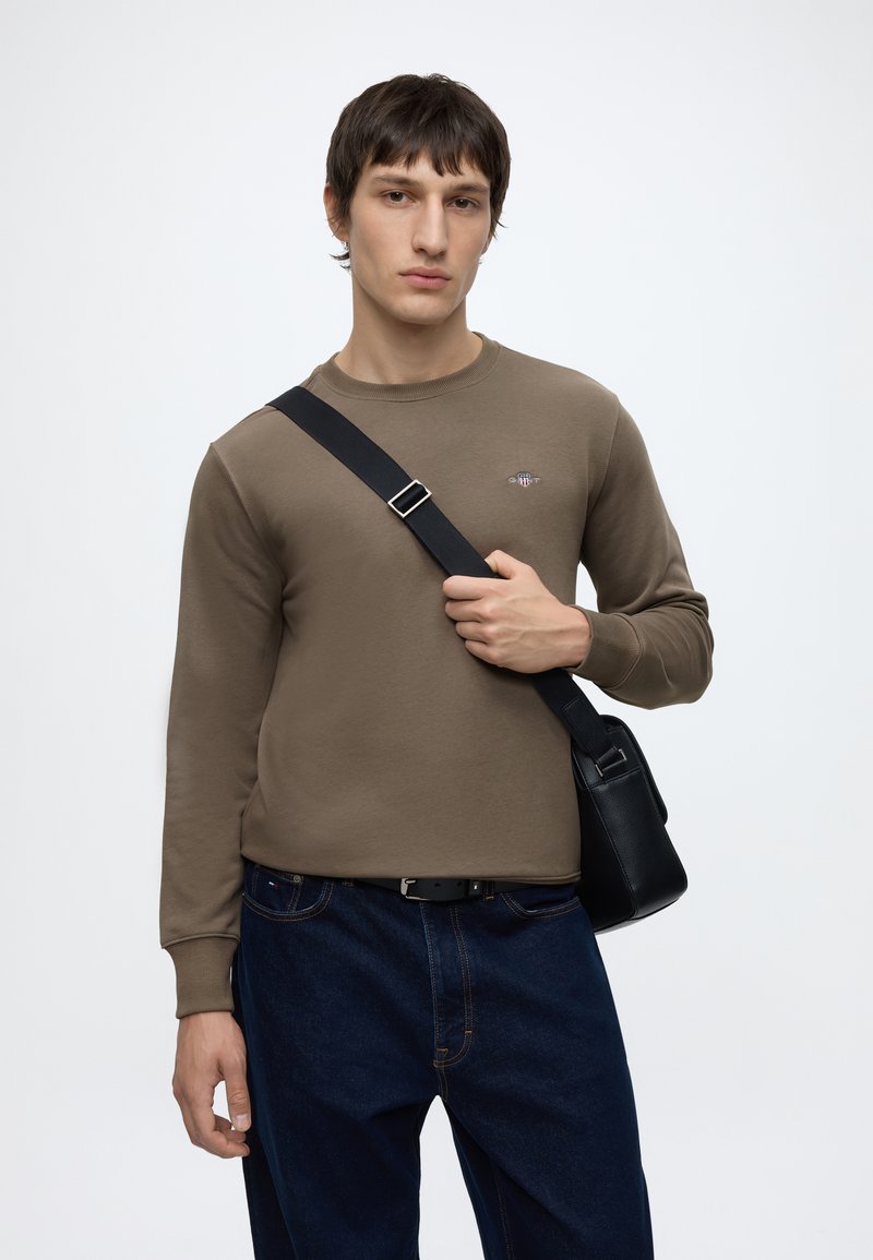 En ung mand iført en brun sweatshirt og mørke jeans holder en sort crossbody-taskerem over skulderen mod en ensfarvet baggrund.