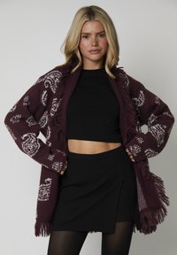 Cardigan de punto burdeos con patrones de paisley blancos y detalles de flecos. Llevado abierto, sobre un top corto negro y una falda negra.
