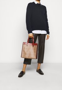 Tote taske med tan mønster og burgunder håndtag. Glat tekstur, struktureret form og forstærkede hjørner. Sammen med en mørk sweater og læderbukser.