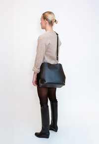 Γυναίκα με ξανθά μαλλιά μαζεμένα σε κότσο, φοράει μπεζ πουλόβερ, μαύρο καλσόν, ψηλές μαύρες μπότες και κρατά μια μεγάλη μαύρη τσάντα ώμου.