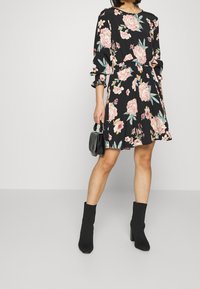 Kvinna i en svart blommig klänning med långa ärmar, som håller en liten svart handväska och har på sig svarta ankelboots med blockklackar.