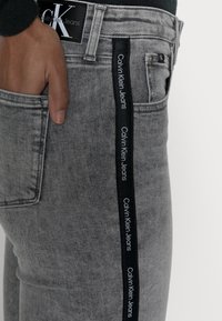 Grå denimjeans med sidelister som har "Calvin Klein Jeans"-logotyp. Materialet har en slät textur och en figurnära design.