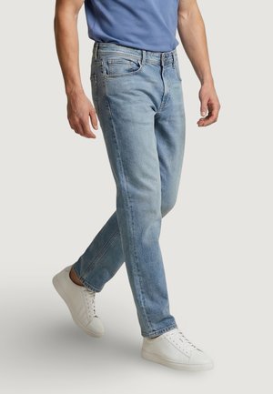 Hombre caminando, vestido con vaqueros azul claro, zapatillas blancas y una camiseta azul sobre un fondo liso.