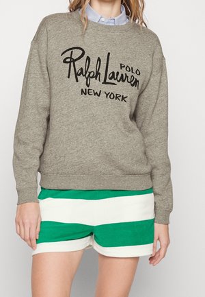 Kvinde iført grå Ralph Lauren Polo New York sweatshirt og grønne og hvide horisontalt stribede shorts, stående mod hvid baggrund.
