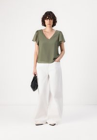 Oliven Grøn V-hals bluse med korte flutterærmer, parret med brede hvide bukser. Modellen holder en sort, struktureret håndtaske.