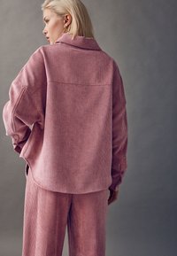 Haut en velours côtelé rose avec col, manches larges et coupe décontractée ; associé à un pantalon assorti, présentant une texture côtelée verticale.