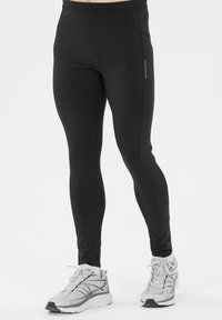 Leggings atléticos negros, con un diseño ajustado, textura suave y detalle de logo reflectante. Combinados con zapatillas de correr grises.