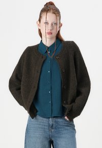 Dark brown knitted cardigan with a round neck, featuring six black buttons and wide sleeves, worn over a teal button-up shirt and blue jeans.