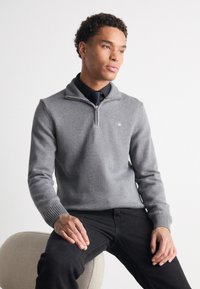Uomo che indossa un maglione grigio con zip a quattro quarti sopra una camicia nera con cravatta, seduto su uno sgabello beige chiaro e arrotondato su sfondo bianco.