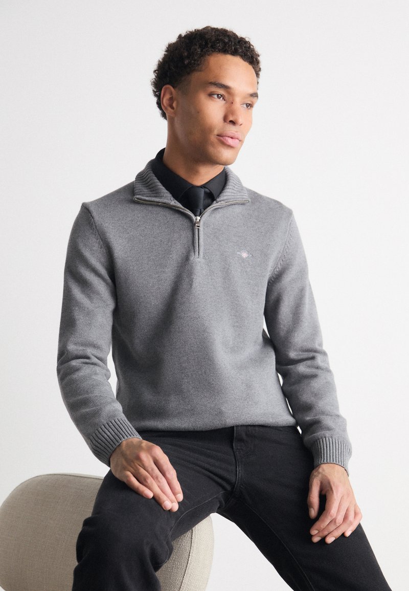 Grauer Pullover mit Reißverschluss und weichem Strickmaterial, mit gerippten Bündchen. Verfügt über ein dezentes Logo auf der Brust und wird mit schwarzen Hosen kombiniert.