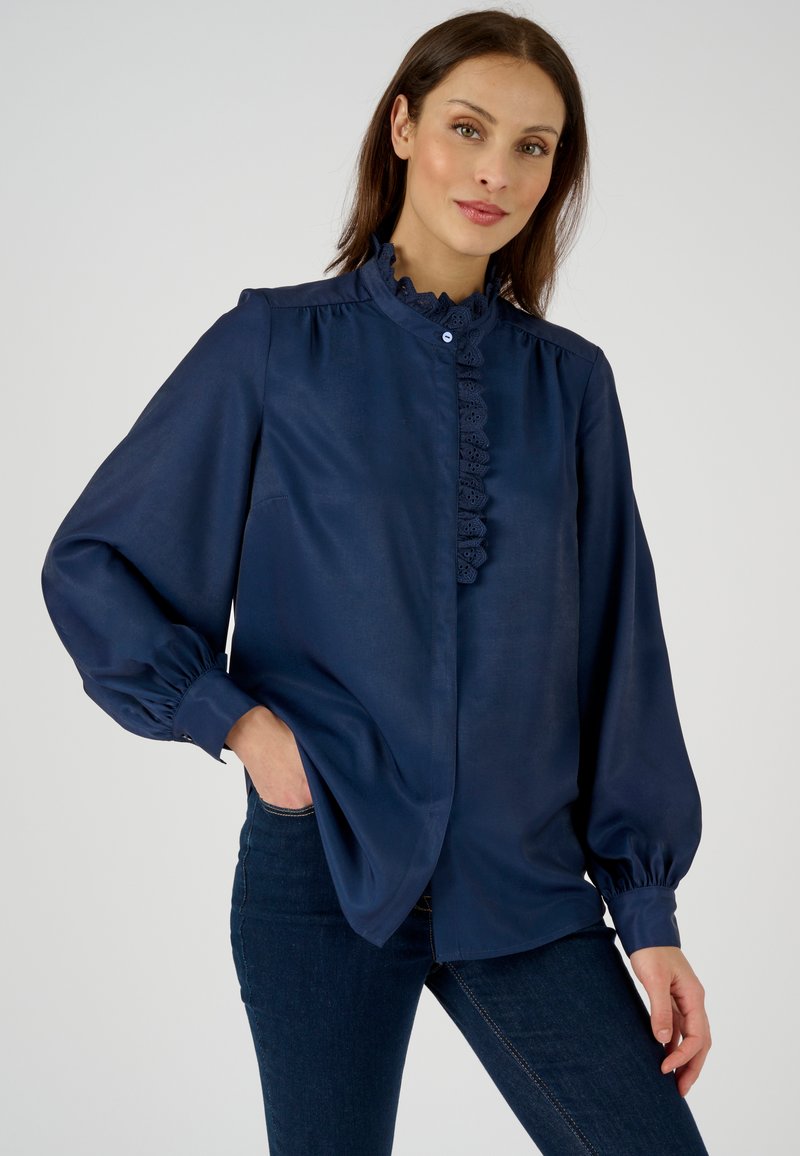 Chemisier marine avec col volanté, manches longues bouffantes et fermeture à boutons. Fabriqué en tissu lisse, il présente de délicates accents en dentelle.