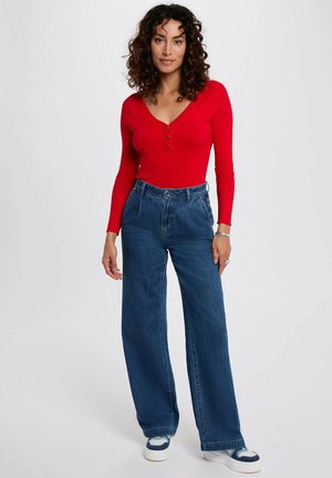 Top a maniche lunghe rosso a coste con scollatura a V e dettaglio di bottoni, abbinato a jeans di denim blu a vita alta e gamba larga. Le sneakers completano l'outfit.