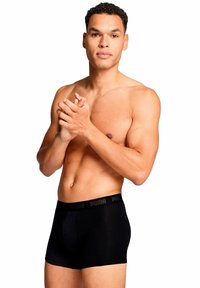 Calzoncillos boxers negros hechos de una tela suave con una cintura elástica que presenta un logotipo negro. El modelo tiene un físico tonificado y está sin camiseta.