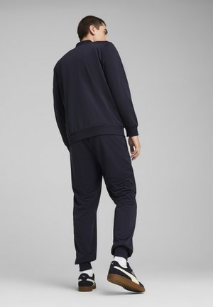 Navy trainingspak met een ritsjack en toelopende broek, beide gemaakt van gladde stof. Gecombineerd met zwart en crèmekleurige sneakers.