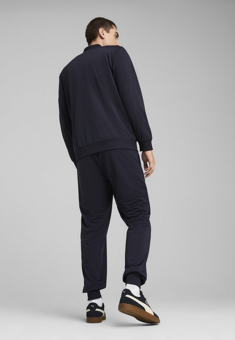 Navy trainingspak met een ritsjack en toelopende broek, beide gemaakt van gladde stof. Gecombineerd met zwart en crèmekleurige sneakers.