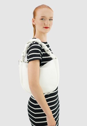 Witte handtas met een gestructureerde afwerking en een dikke, gevlochten handgreep. Model draagt een zwart-wit gestreepte jurk en toont het ontwerp van de tas.