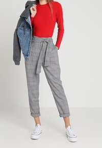 Donna che indossa un top rossi a coste, pantaloni a quadri a vita alta con cintura, sneakers bianche, mentre tiene una giacca grigia e in denim su una spalla.