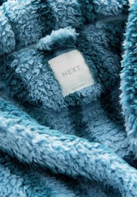 Weiche blaue Fleece-Decke mit einer strukturierten, flauschigen Oberfläche. Mit einem Etikett, auf dem "NEXT" und "Hergestellt in Kambodscha" steht.