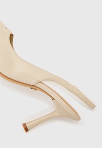 Sandalo con tacco alto beige, con tomaia in pelle liscia, lati aperti e tacco sottile. Presenta una forma curva e una piccola punta del tacco trasparente.