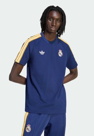 Person mit dunkler Haut und Dreadlocks, trägt ein blaues Adidas Real Madrid Trikot und passende Hose mit gelben Streifen, Hände hinter dem Rücken.