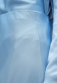 Svetlo modra bluza z teksturirano, gladko tkanino. Ima kroj po meri, detajle na stranskih šivih in pleteni rokav na manšeti v nekoliko temnejši modri barvi.