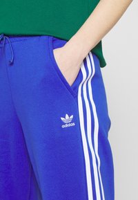 Modré tepláky se třemi bílými pruhy po stranách a vyšitým logem Adidas na stehně. Vyrobené z měkké látky.