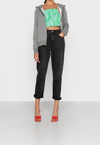 Grijze hoodie met ritssluiting over een groen-wit patroon crop top, gecombineerd met zwarte opgerolde jeans en doorzichtige rode sandalen met hak.