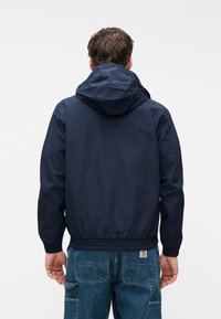 Άνδρας με μπλε σκούρο (ναυτικό) μπουφάν με κουκούλα και μπλε τζιν, εικονισμένος από πίσω, σε απλό λευκό φόντο.