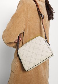 Bolso de cuero color beige tipo bandolera con un patrón geométrico en relieve, costuras en contraste y correa ajustable, colocado sobre una chaqueta de pana texturizada.