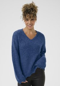 Blauer, strukturierter Strickpullover mit V-Ausschnitt, überlängten Schultern und asymmetrischem Saum. Weiches Material und lockere Passform für hohen Tragekomfort.