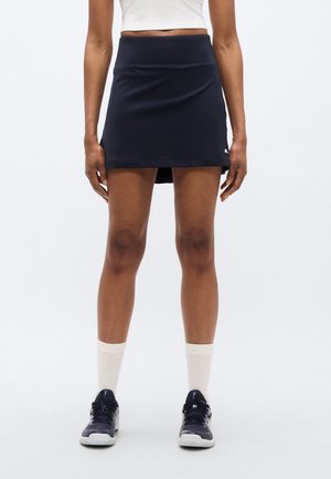 Jupe de sport bleu marine avec une taille haute, une texture lisse, longueur mi-cuisse et des fentes latérales pour faciliter les mouvements, assortie à des baskets grises.