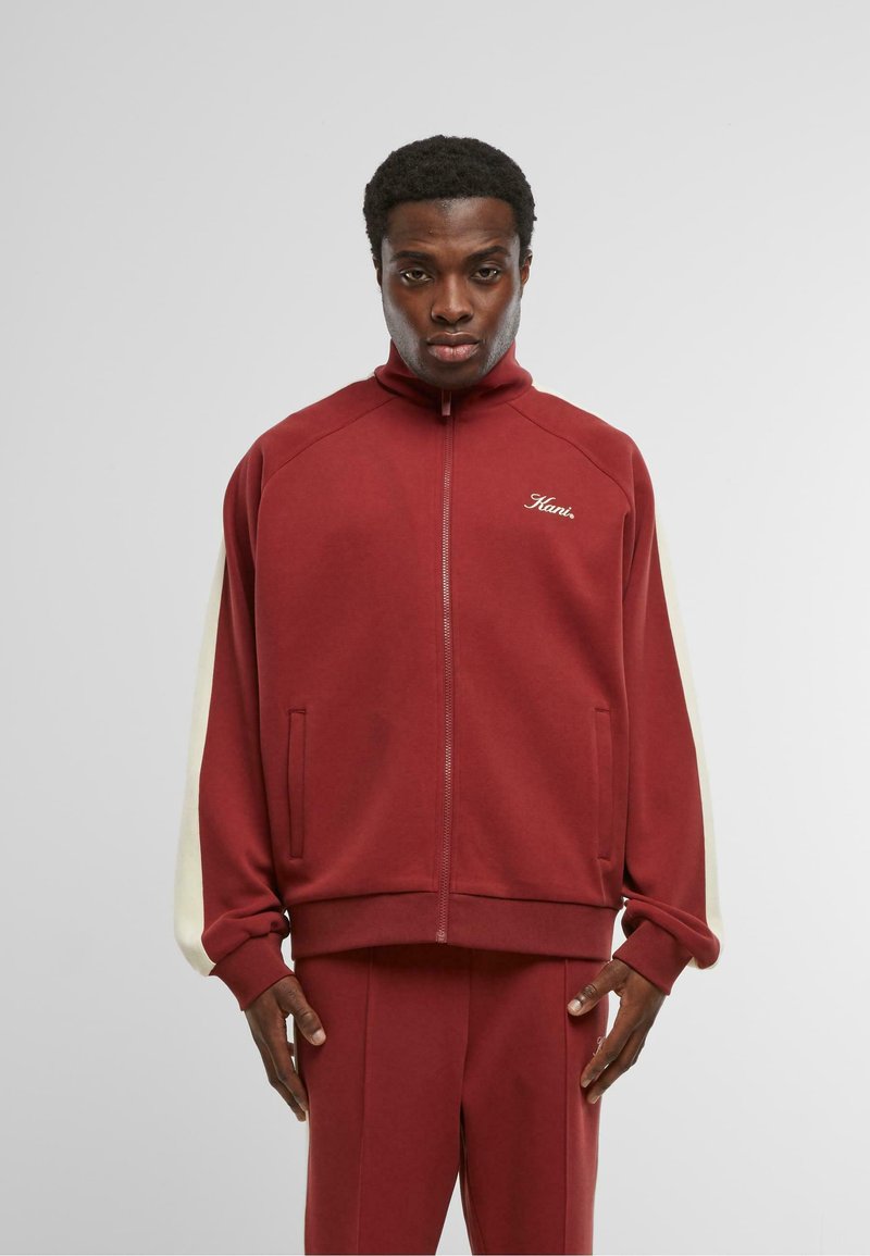 Muž v červenej zipsovej športovej bunde a zodpovedajúcich nohaviciach s bielymi pruhmi na rukávoch, stojí proti jednoduchému šedému pozadiu.