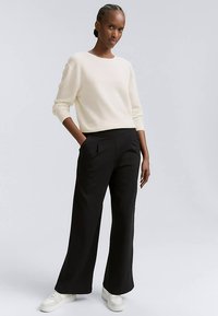 Krämfärgad stickad tröja med långa ärmar kombinerad med svart vida byxor med hög midja; modellen bär vita sneakers. Mjuk textur, avslappnad passform.