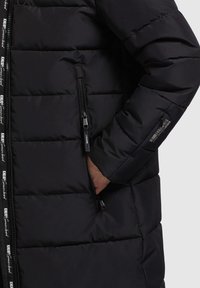 Zwarte pufferjas met een gequilt ontwerp, voorzien van een ritszak aan de zijkant en merkritsen. Glad, glanzende textuur.