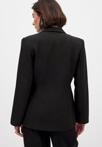 Melnā pieguļošā žakete ar gludu tekstūru, ar nogrieztām atsegumiem un garām piedurknēm. Aizmugurē ir šūtas šuves, kas veido strukturētu siluetu.