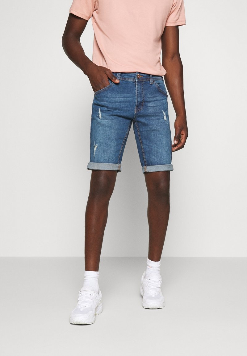 Shorts en denim bleu clair avec une finition délavée et des détails usés, présentant des ourlets roulés et une fermeture à bouton. Assortis avec des baskets blanches.