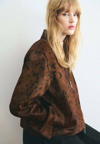Veste bomber marron à pois fabriquée en tissu textur�é, avec une fermeture éclair sur le devant et une coupe décontractée. Les détails incluent un col et des poignets côtelés.