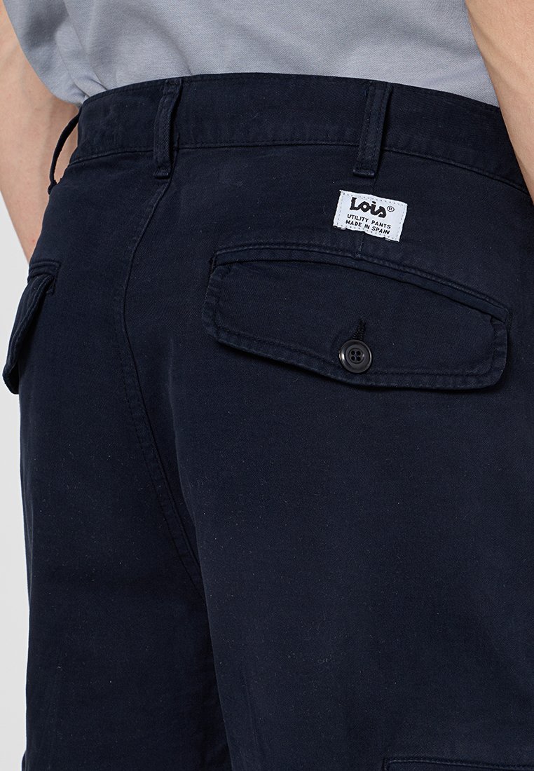 Granatowe spodnie robocze o gładkiej fakturze, z tylnymi kieszeniami zapinanymi na guziki i metką z napisem "Lola Spodnie Robocze Wyprodukowano w Hiszpanii."