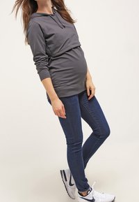 Felpa grigia con tasca a marsupio, maniche lunghe e cappuccio con cordino, abbinata a jeans skinny blu scuro e scarpe sportive bianche.