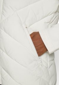 Main à la peau foncée dans la poche avant zippée d'une veste blanche matelassée avec des coutures diagonales.