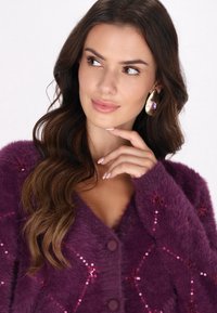 Cárdigan morado suave con botones redondos, decorado con brillantes lentejuelas rojas en un patrón de diamantes. Pendientes grandes de oro con una piedra morada.