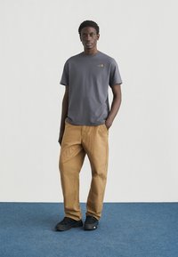 Γκρι κοντομάνικο μπλουζάκι με λογότυπο στο στήθος, σε συνδυασμό με ανοιχτό καφέ υφασμάτινο παντελόνι και μαύρα αθλητικά παπούτσια σε ουδέτερο φόντο.