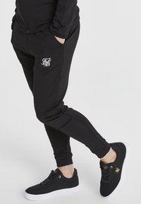 Schwarze Jogginghosen mit schlanker Passform, die ein weißes gesticktes Logo auf dem Oberschenkel tragen. Kombiniert mit schwarzen Sneakers mit weißen Sohlen und goldenen Akzenten.
