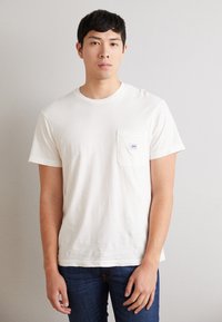Weißes Baumwoll-T-Shirt mit kurzen Ärmeln, rundem Halsausschnitt und einer linken Brusttasche, die ein kleines Markenlabel trägt. Getragen mit dunkelblauen Jeans.