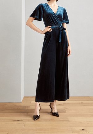 Marineblauwe velvet jumpsuit met brede korte mouwen, V-hals en stoffen ceintuur in de taille, gecombineerd met zwarte puntige hakken op een houten vloer.