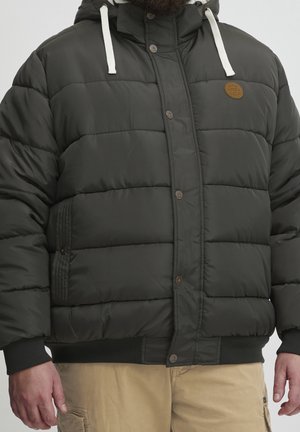 Man wearing dark green padded jacket with white drawstrings and beige pants, standing with hands relaxed at sides.